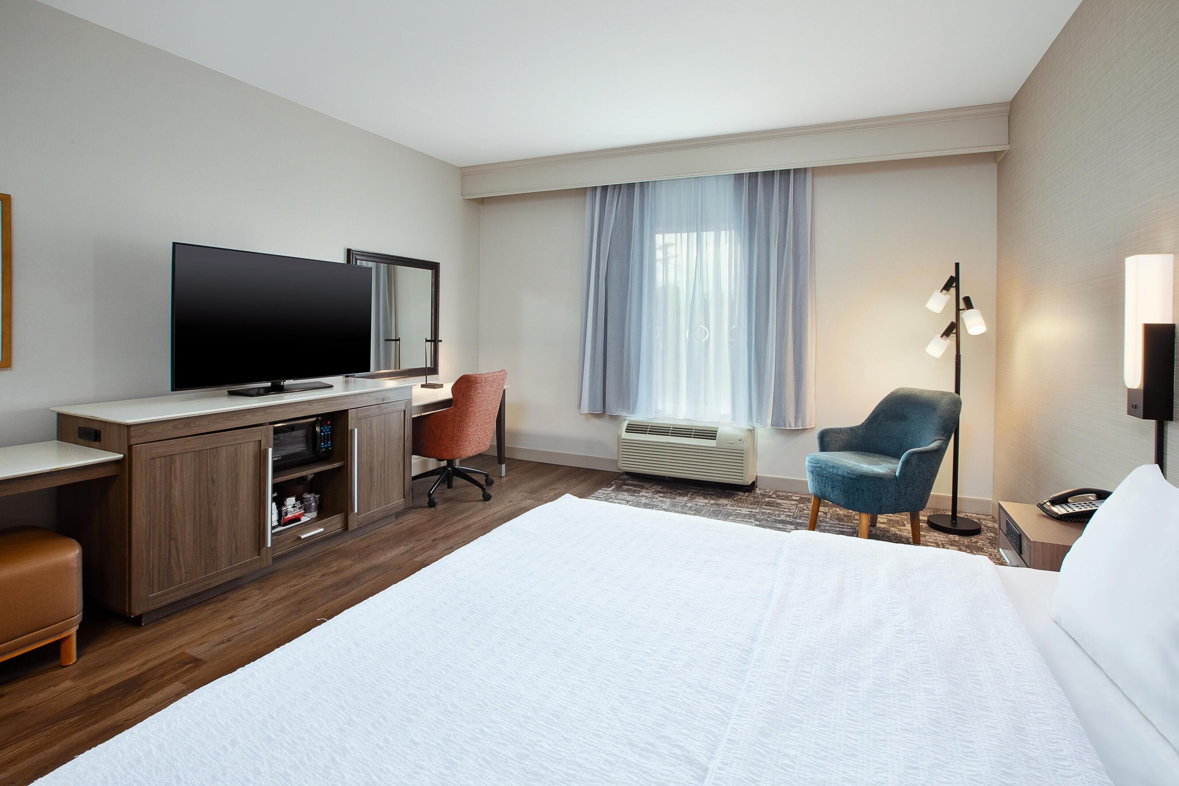 Une chambre d’hôtel avec un grand lit, un bureau doté d’un téléviseur à écran plat, une chaise, un lampadaire et une fenêtre avec des rideaux.