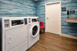 A laundry room with a front-loading washer and dryer, a red chair, and a door with a sign indicating emergency contact numbers. - Hampton Inn North Little Rock McCain Mall (North Little Rock)