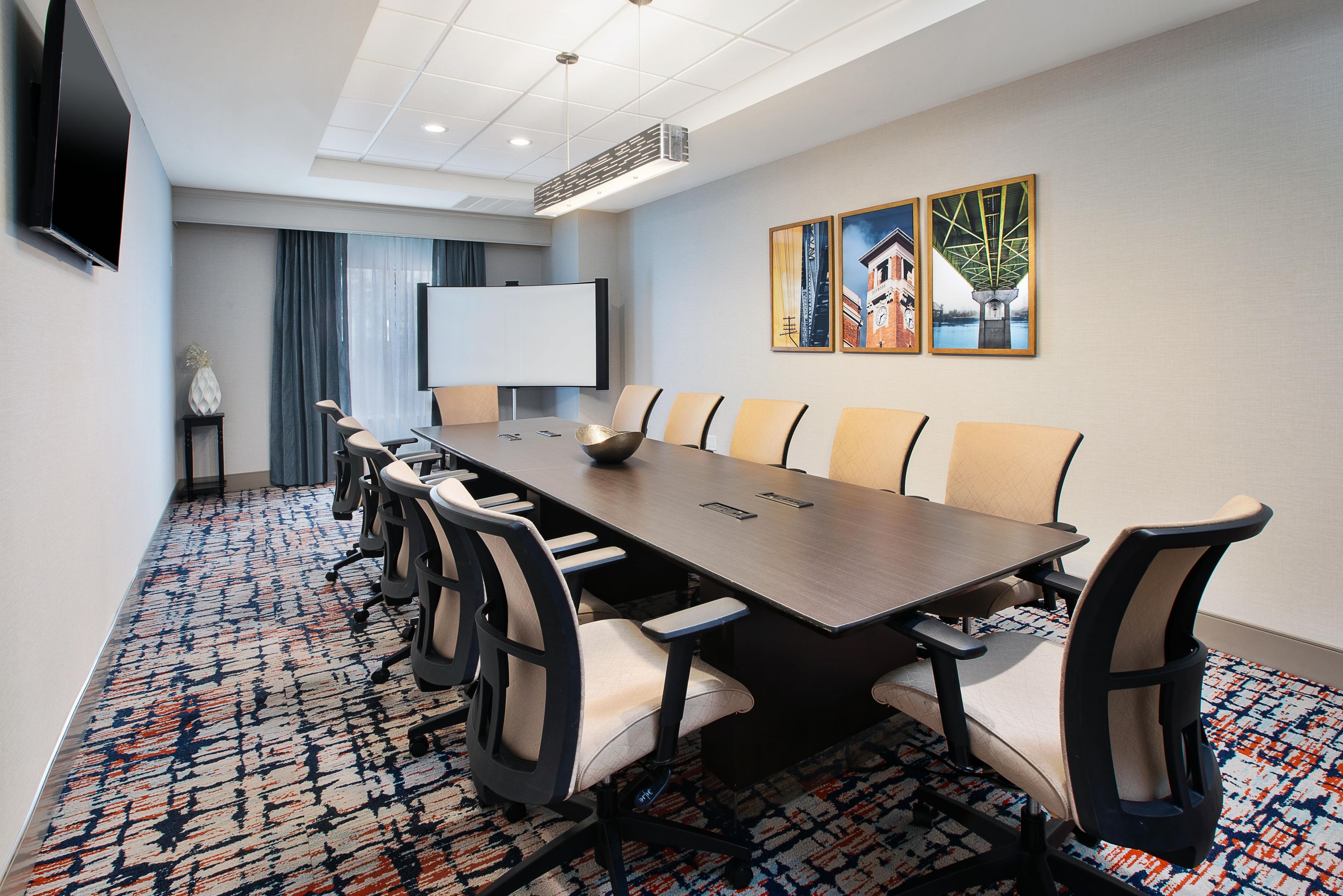 A conference room with a long table, chairs, a projector screen, and artwork on the wall.