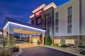Un hotel Hampton Inn con fachada moderna, acceso cubierto y un muro decorativo de piedra.