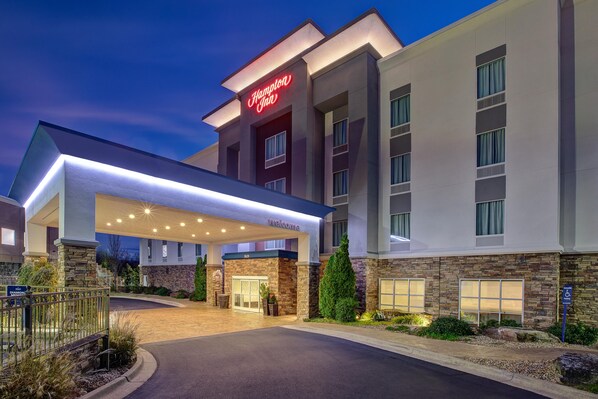 Un hotel Hampton Inn con fachada moderna, acceso cubierto y un muro decorativo de piedra.
