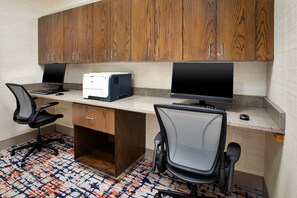 A workspace with two desks, each with a computer monitor, keyboard, and mouse. There are two office chairs. The room has wooden cabinets above the desks and a patterned carpet on the floor. - Hampton Inn North Little Rock McCain Mall (North Little Rock)