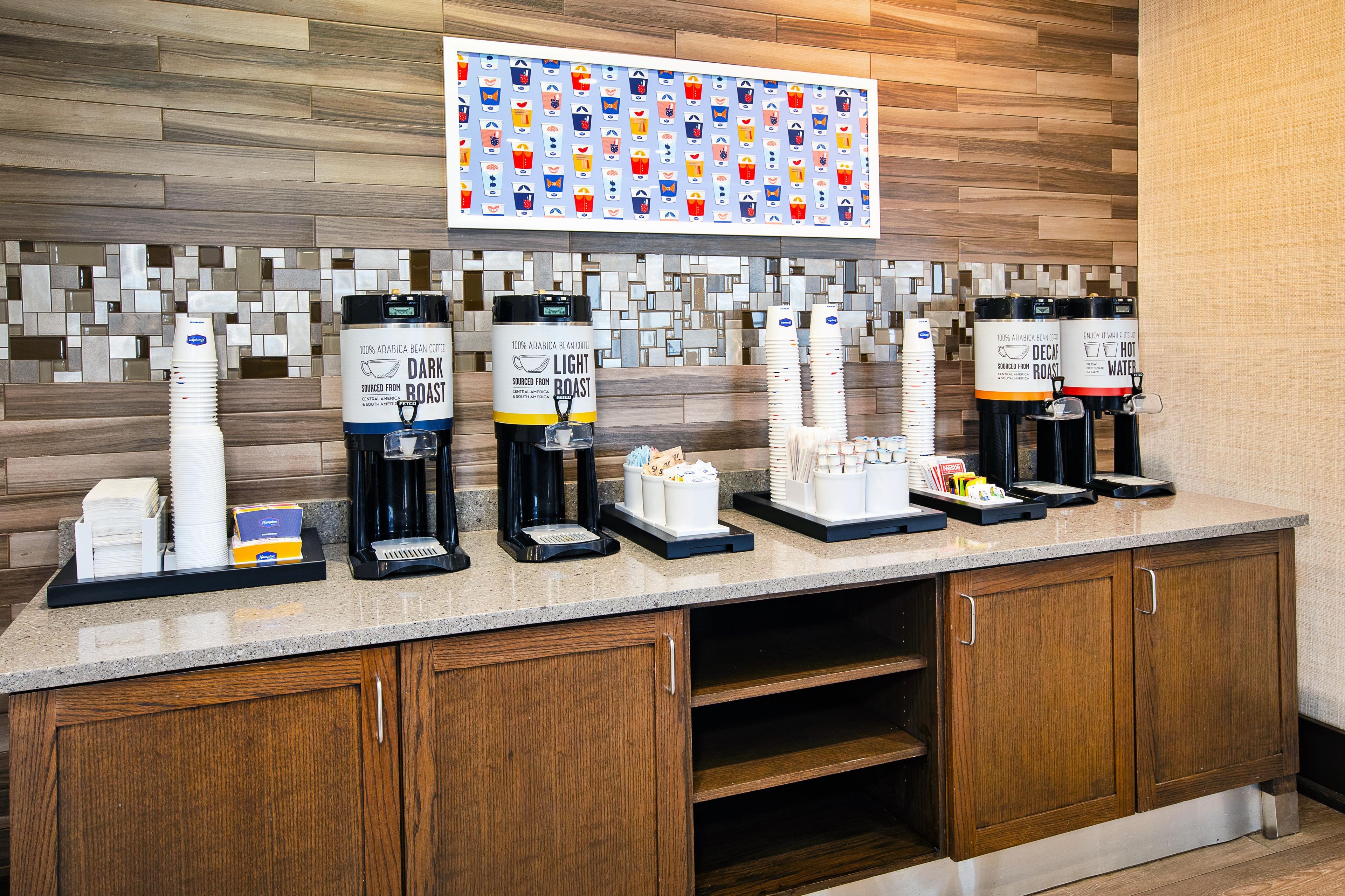 A coffee station with multiple coffee dispensers, disposable cups, and a wall-mounted menu board.