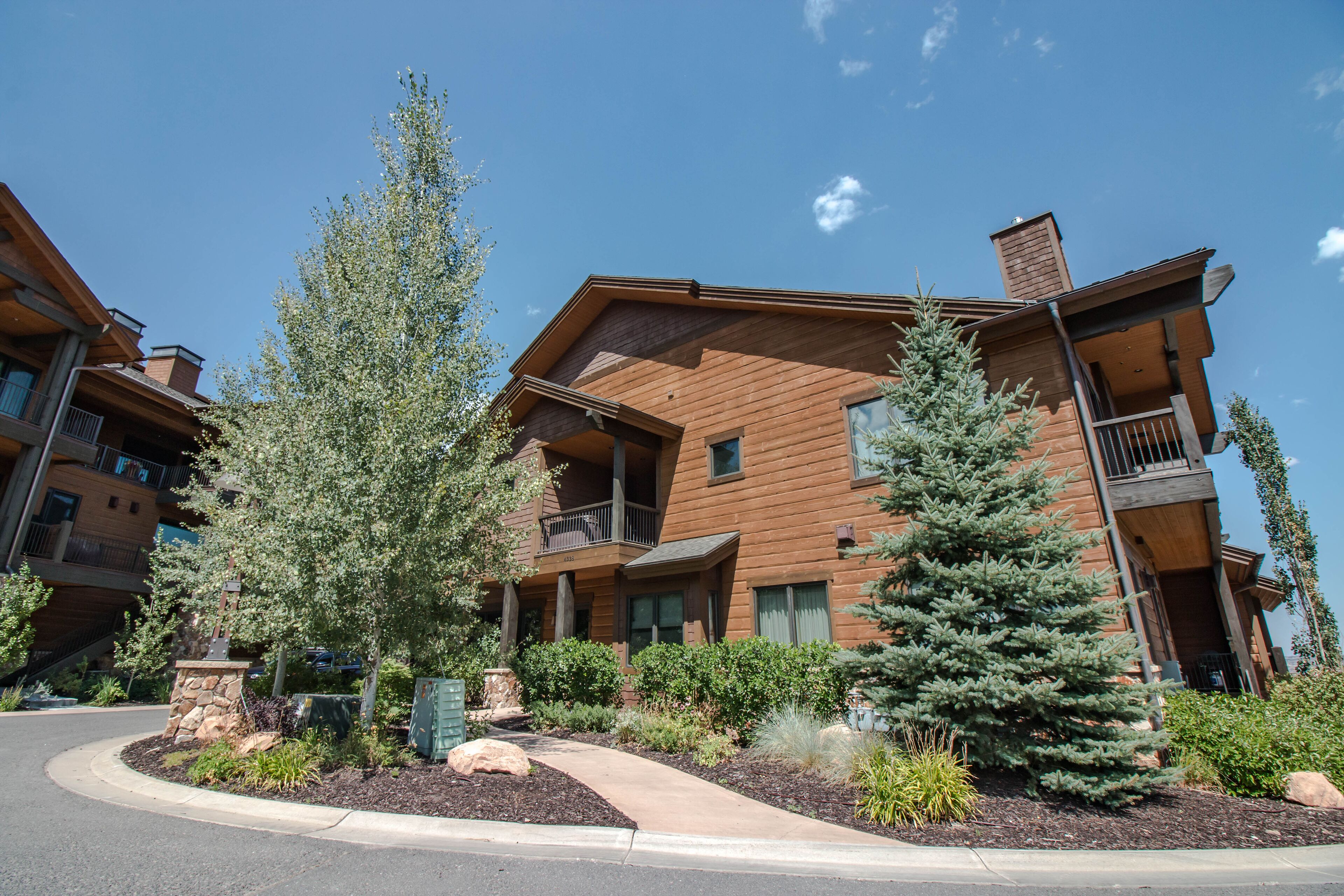 A multi-story building with a wooden exterior, balconies, and a landscaped front yard with trees and shrubs.