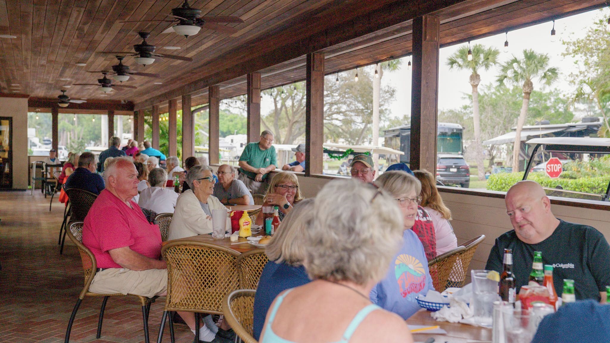 A covered outdoor dining area with wicker chairs and tables, large windows, and a view of palm trees and parked cars.