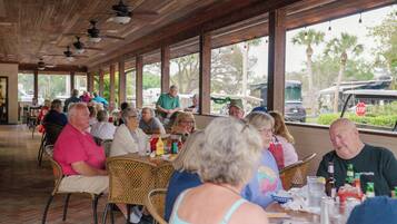 A covered outdoor dining area with wicker chairs and tables, large windows, and a view of palm trees and parked cars.