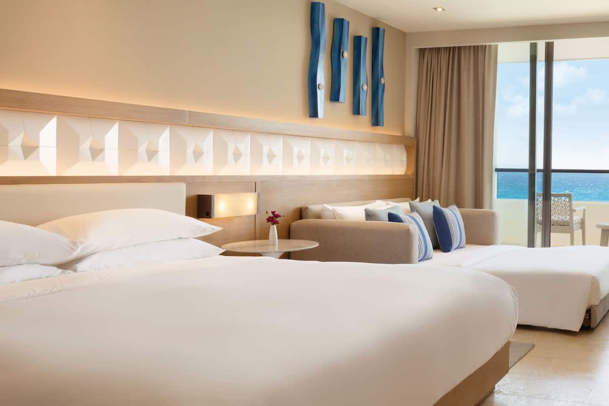 Habitació d'hotel amb dos llits, un sofà, una taula petita amb un ram i vista al mar.