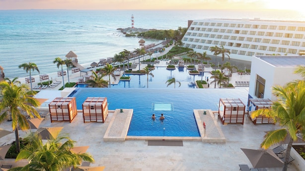 A resort pool with two people swimming, surrounded by palm trees and a view of the ocean. - Hyatt Ziva Cancun All Inclusive (Cancun)