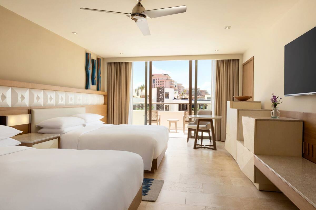Habitació d'hotel amb dos llits, un escriptori, una cadira, un televisor i un balcó amb vistes.