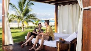A wooden deck with a bed, two people sitting and talking, palm trees, and a view of the beach and ocean. - Hyatt Ziva Cancun All Inclusive (Cancun)