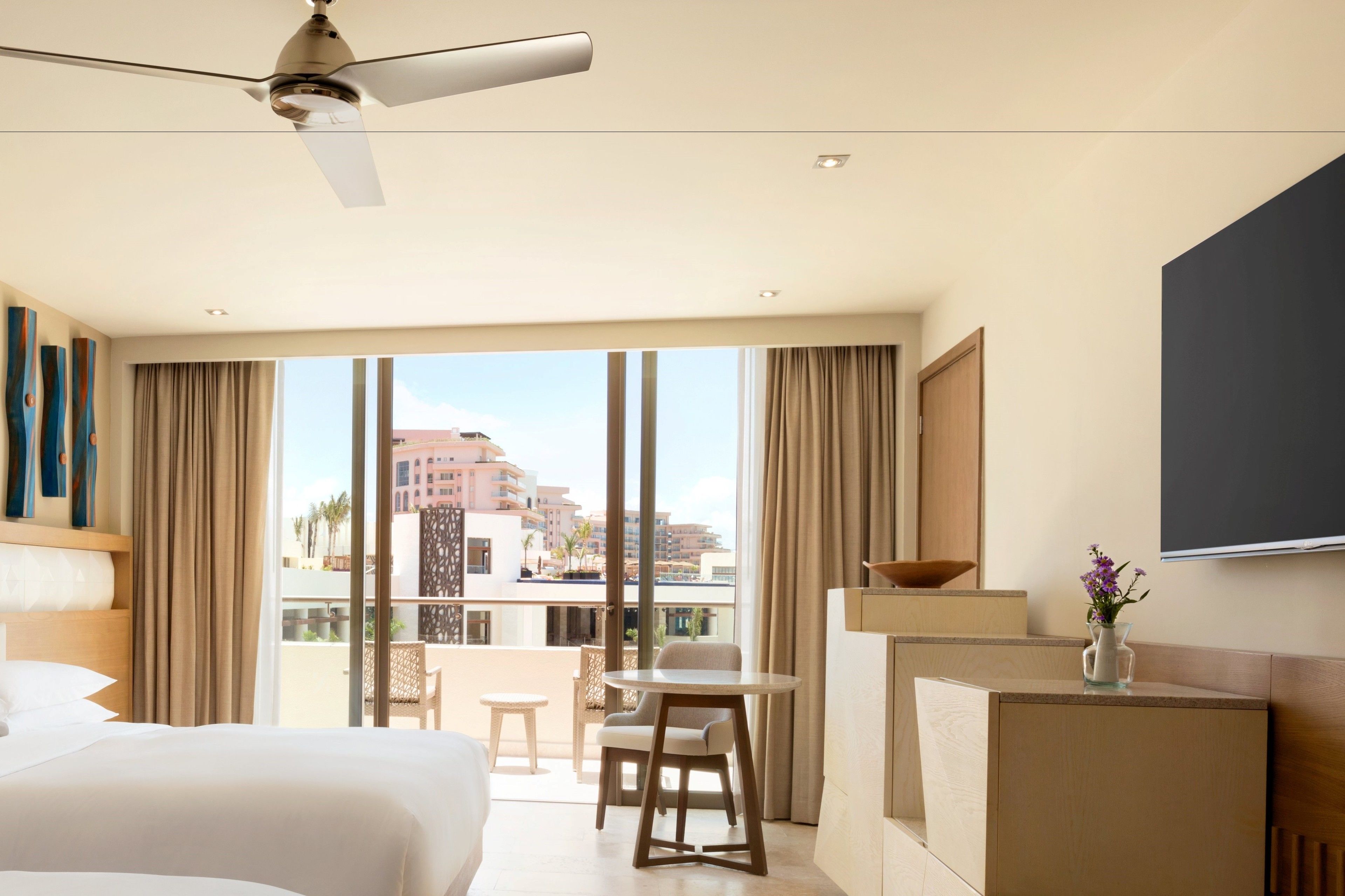 A hotel room with a bed, a desk, a chair, a small table, a ceiling fan, and a large window with a view of buildings.
