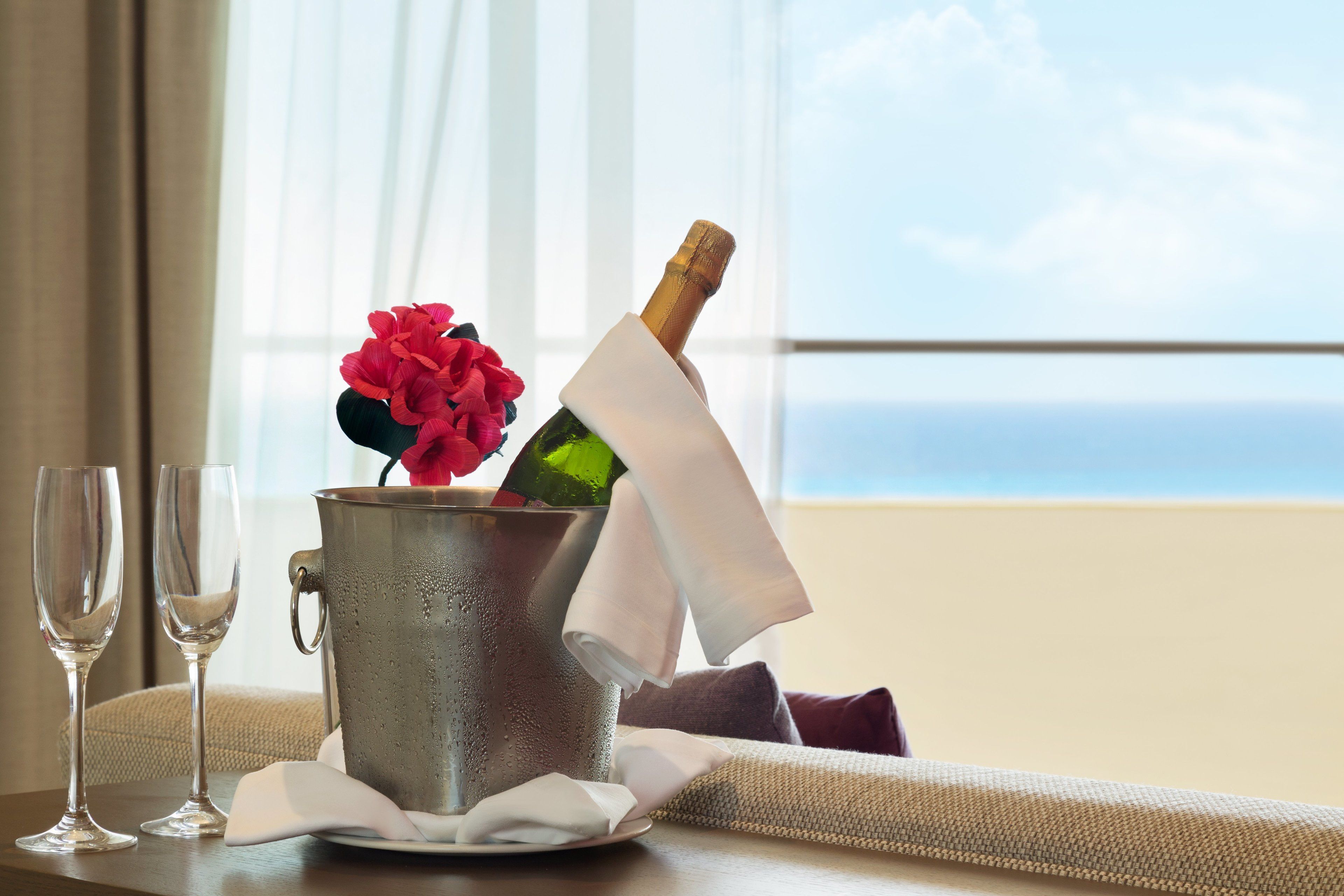 Una botella de champán en un cubo metálico con una flor, dos copas y una servilleta sobre una mesa junto a una ventana con vistas.