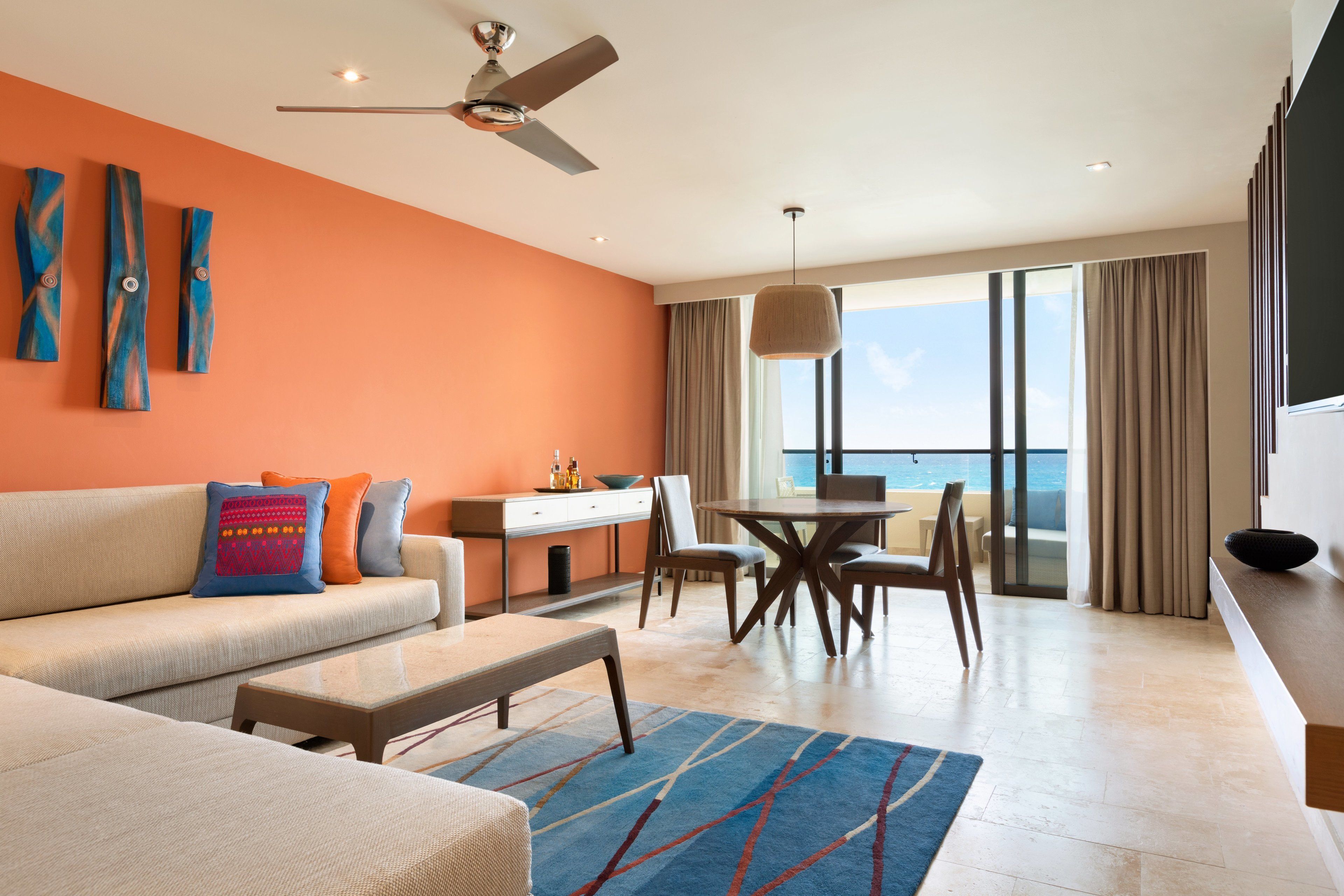 A modern living room with a sofa, coffee table, and dining area. There is a ceiling fan, a large window with a view, and a balcony.