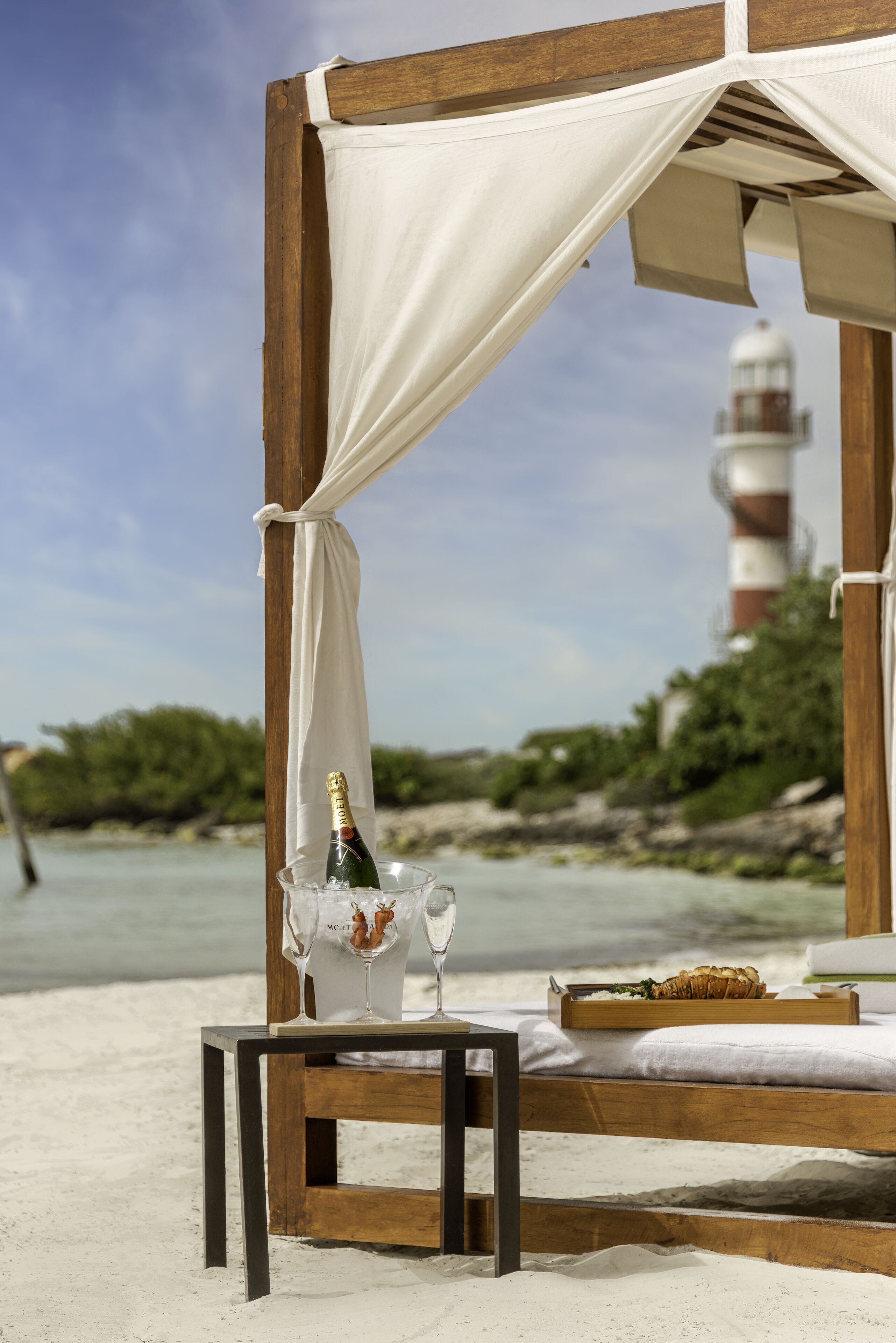 Una cama frente al mar con una botella de champán y copas, una bandeja con comida y una vista de un faro al fondo.