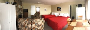 A room with a stone fireplace, two beds with red bedding, a patterned armchair, and a dining table with placemats. - Motel Sainte-Flavie (Sainte-Flavie)