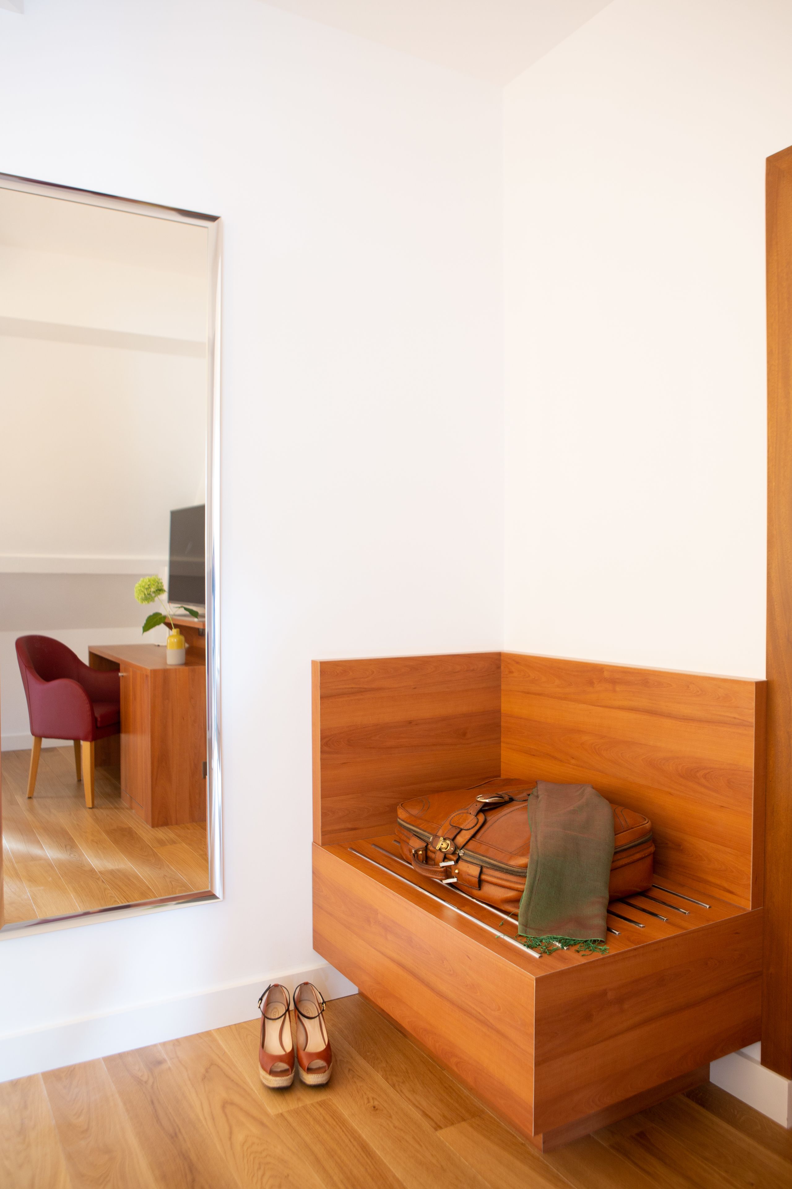 Habitación en esquina con un banco de madera, un par de zapatos, un bolso y un espejo que refleja un escritorio y una silla.