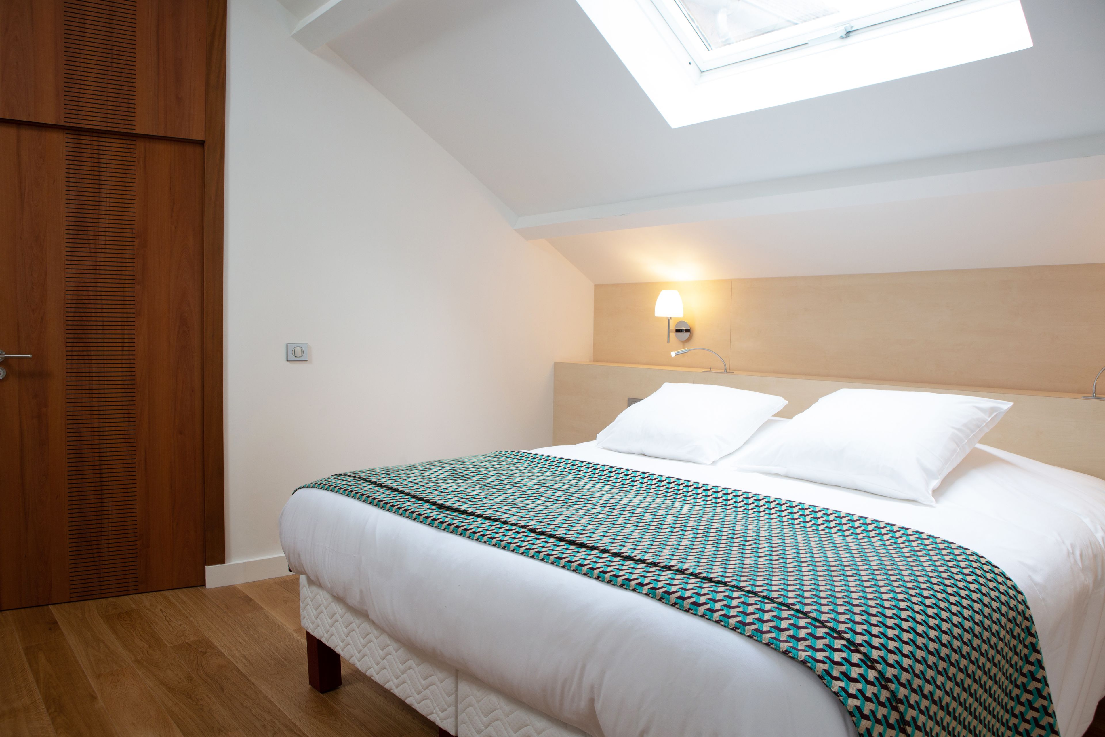 A bedroom with a skylight, a bed with a patterned bedspread, a wooden wardrobe, and a wall-mounted light fixture.