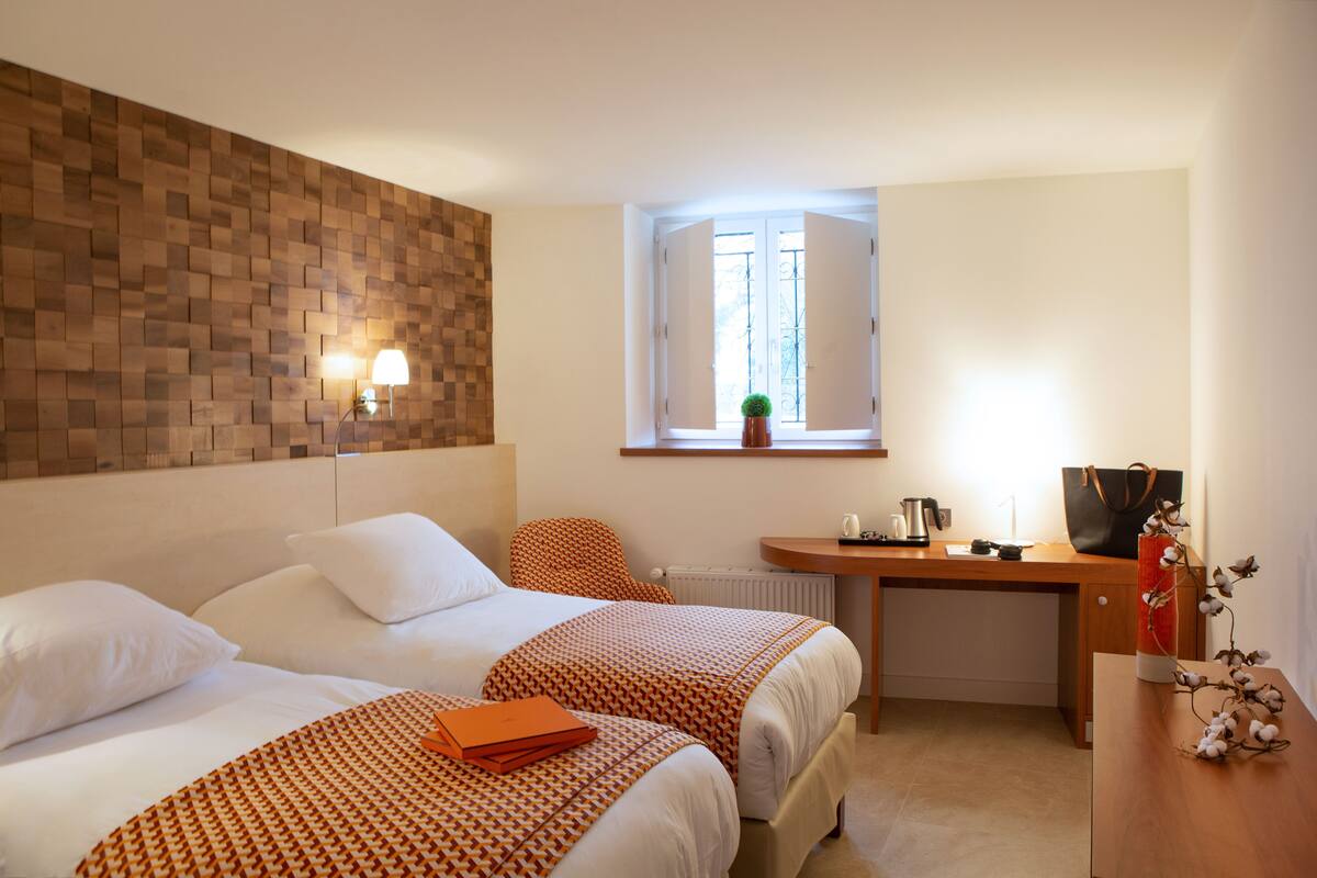 A hotel room with two beds, a wooden headboard, a small table with a kettle, a window with a view, and a plant on the windowsill.