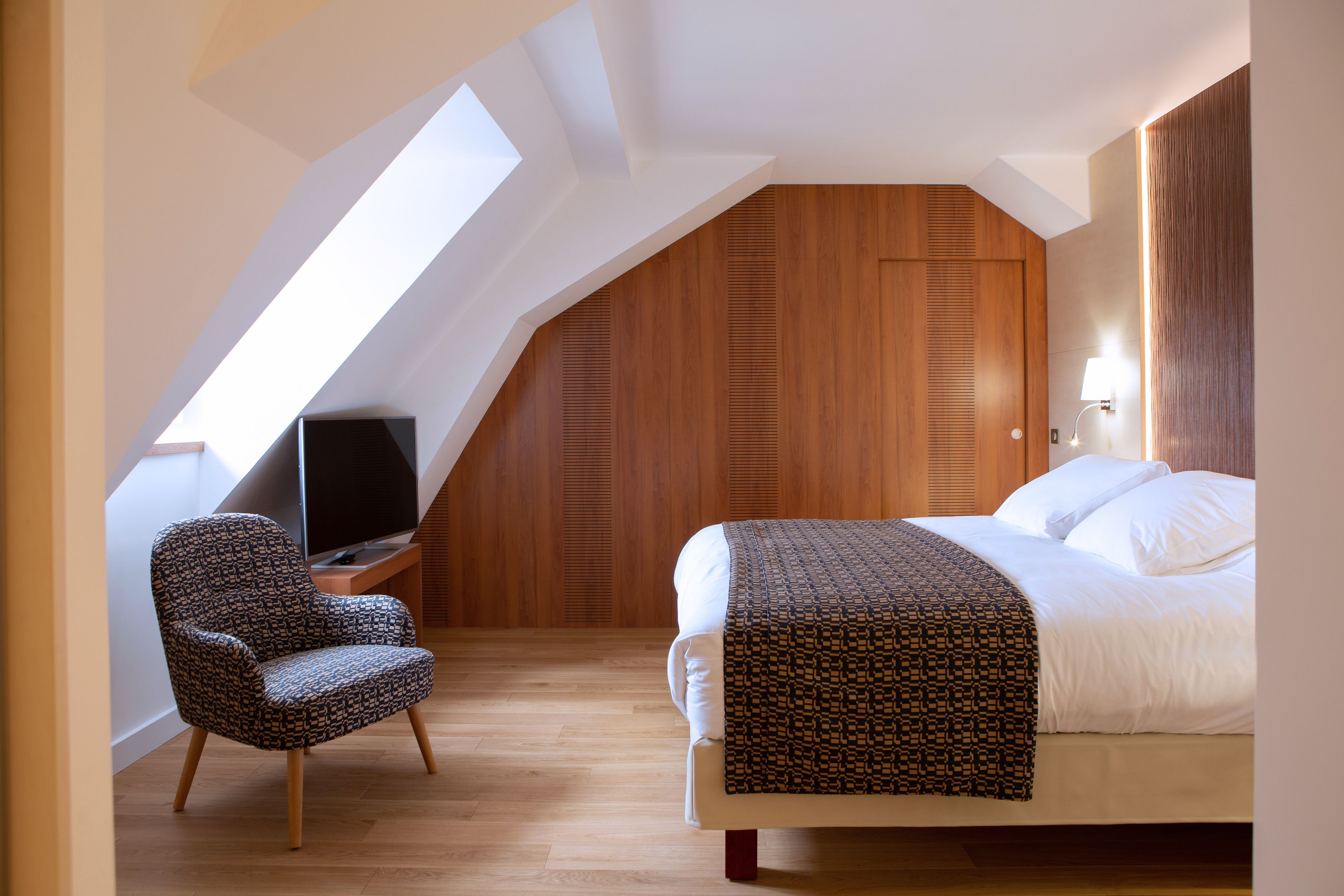Un dormitorio con techo inclinado, una cama con manta estampada, una silla y un televisor sobre un soporte de madera.