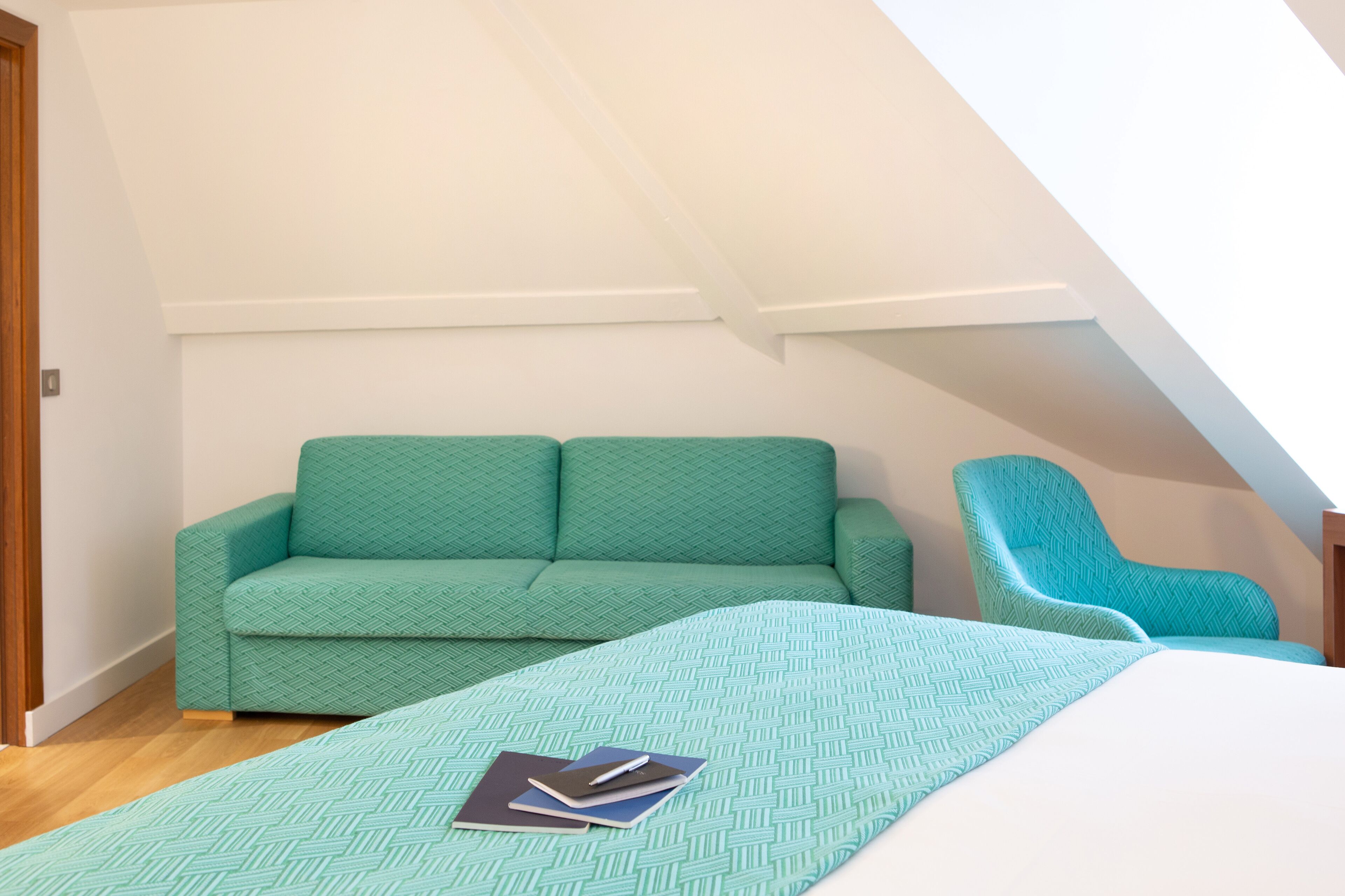 Une pièce avec un canapé vert, un fauteuil turquoise et un lit recouvert d’une couvre-lit à motifs.