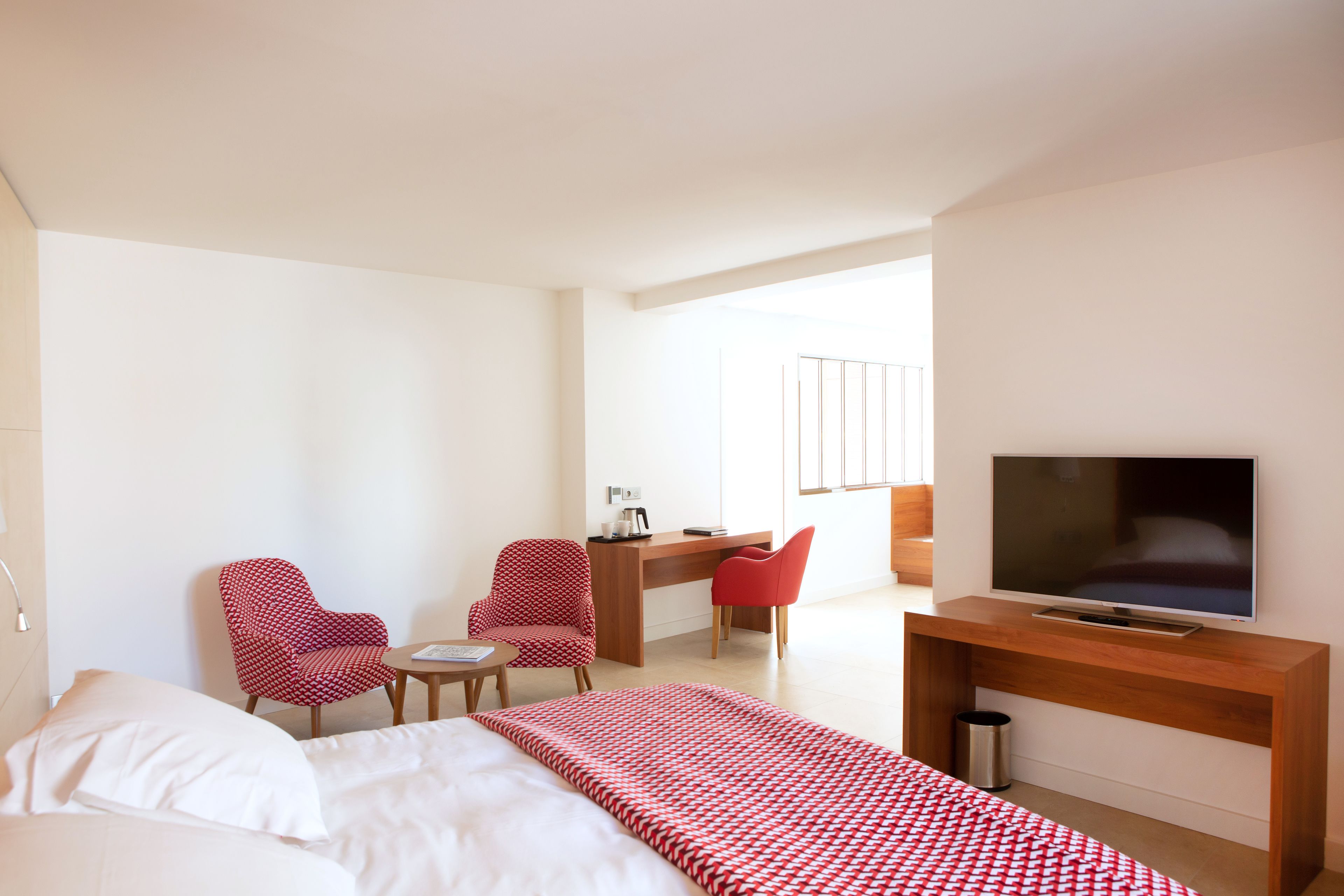 A hotel room with a bed, two red patterned chairs, a desk with a computer, a TV, and a window with a view.