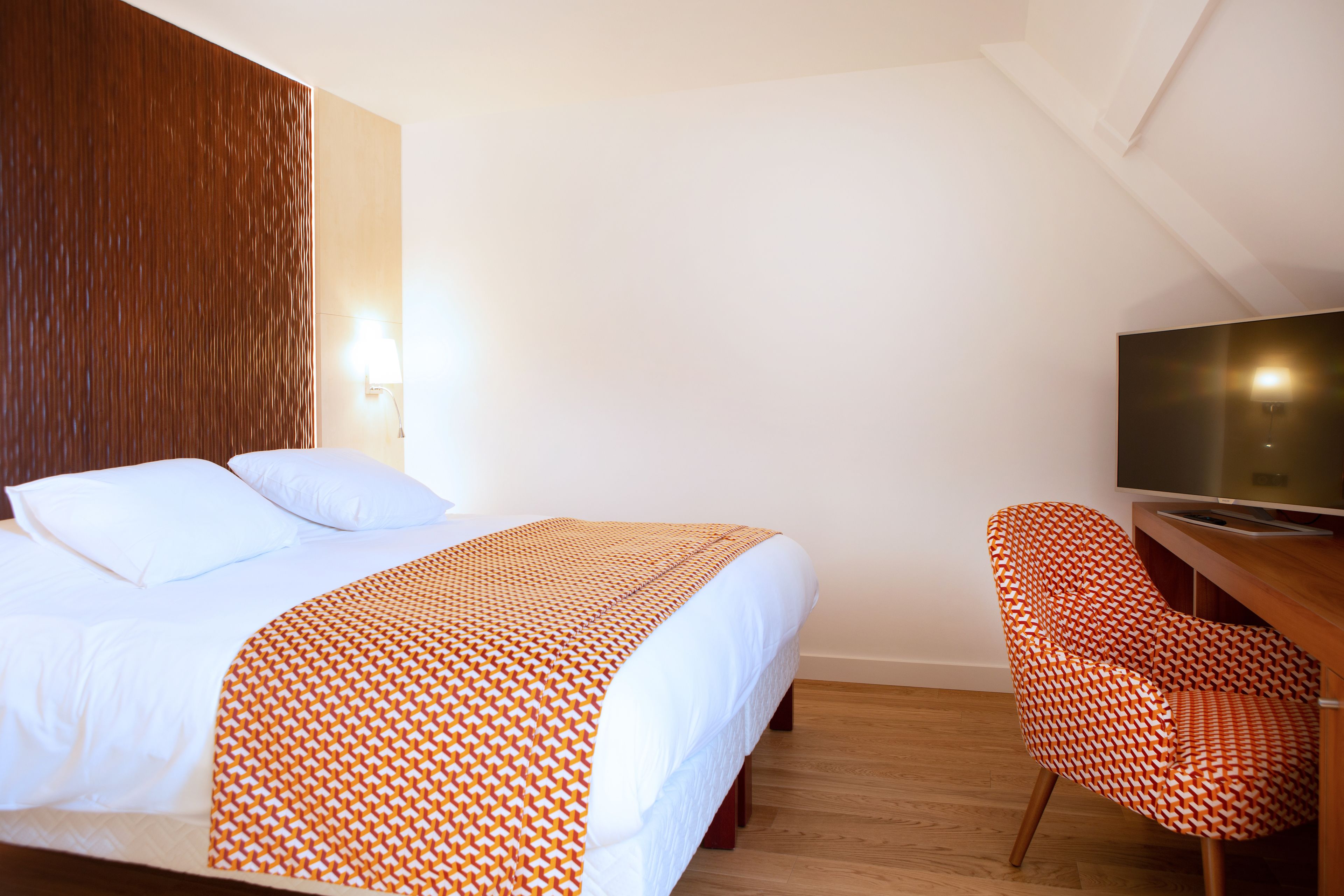 A hotel room with a bed, a TV, a wooden desk, and a patterned chair.