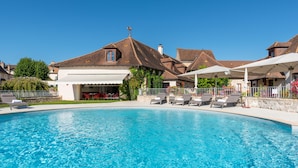Une piscine avec des chaises longues, un patio couvert et un bâtiment doté d’un toit en tuiles.
