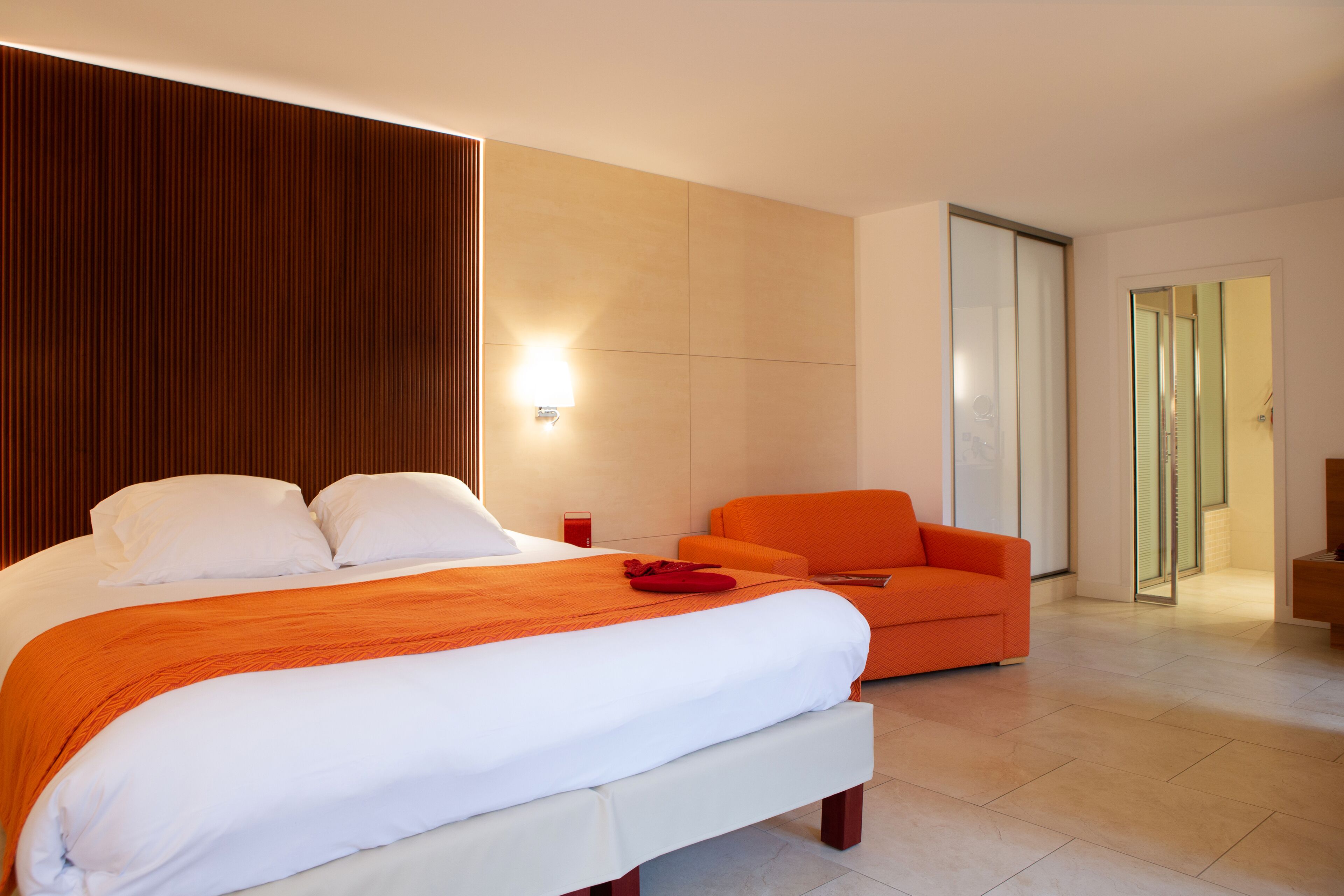 Una habitación de hotel moderna con una cama grande, un sillón naranja y una puerta de cristal que da al baño.