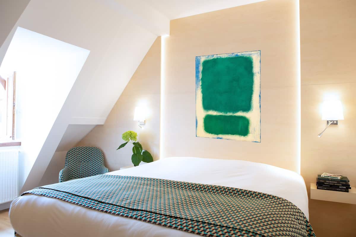 A modern hotel room with a large bed, a green abstract painting on the wall, a potted plant, and a small table with books.