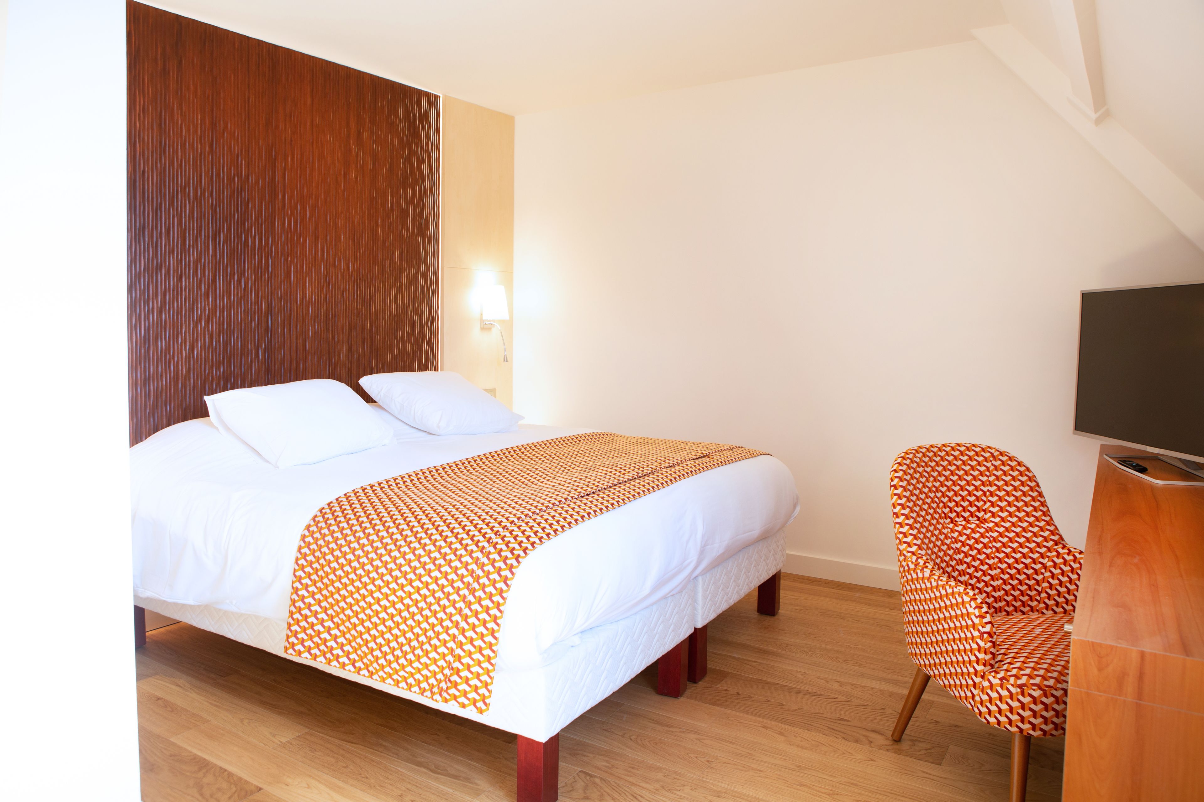 A hotel room with a bed, a chair, a wooden headboard, a wall-mounted TV, and a wooden floor.