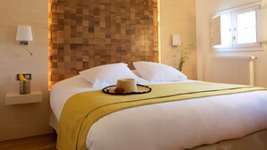 Un lit bien fait, avec du linge de lit blanc, un plaid jaune et un chapeau décoratif posé sur le lit. On y voit une tête de lit en bois, une table de chevet avec une lampe et une fenêtre avec des stores.