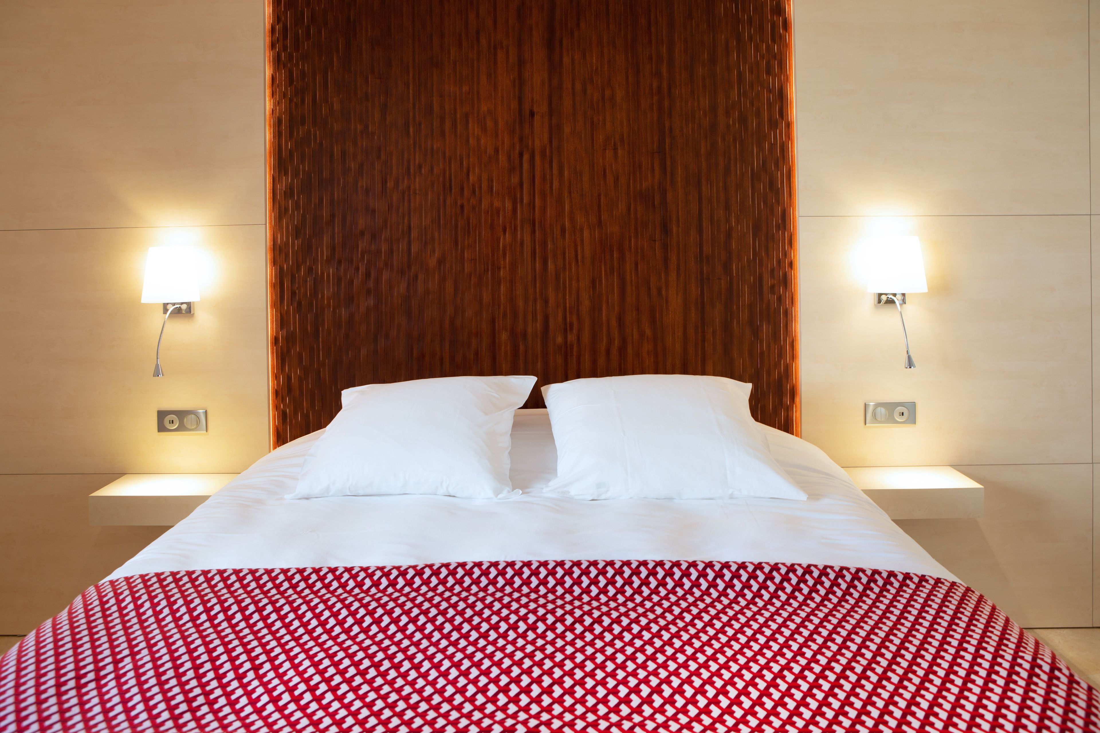 Quarto de hotel com uma cama grande, duas luminárias laterais e cabeceira de madeira.