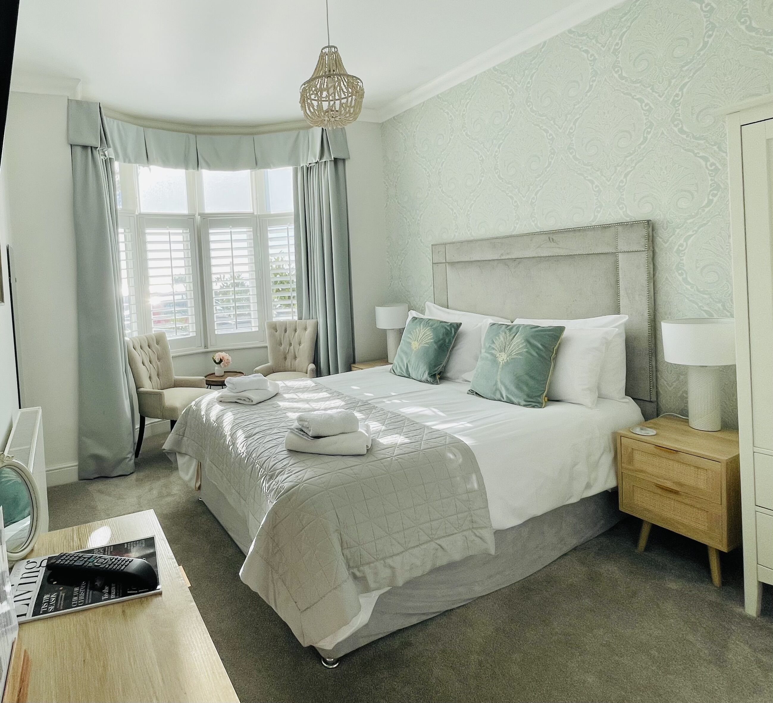 A neatly arranged bedroom with a large bed, a bedside table, a nightstand, a chair, and a window with curtains.