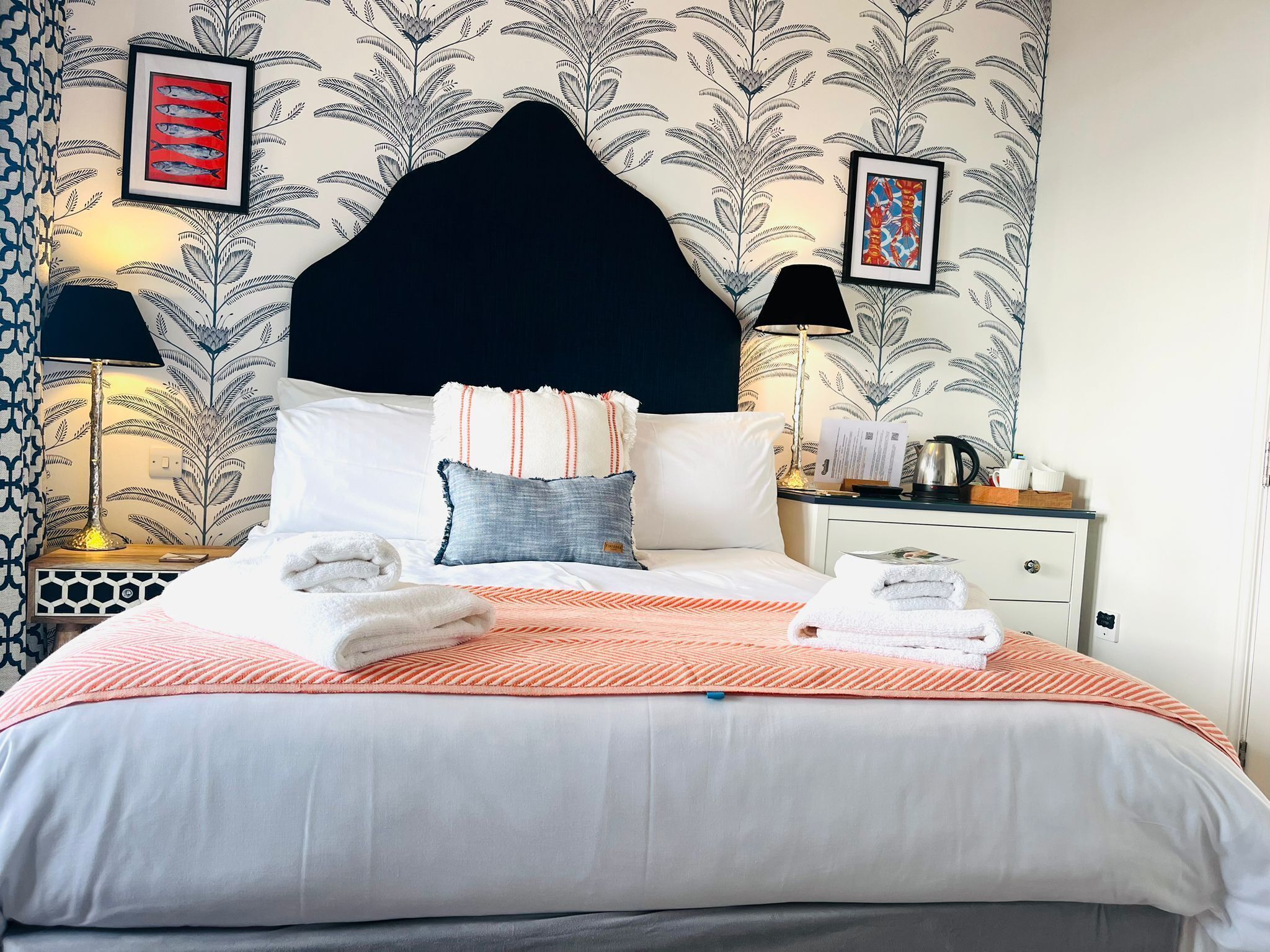 A neatly made bed with a striped blanket, pillows, and folded towels. There are two bedside tables with lamps and a small table with a kettle and cups. The room has a patterned wallpaper and framed artwork on the walls.