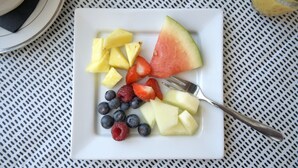 En hvid tallerken med ananas, vandmelon, jordbær, blåbær og hindbær på et bord med et metalmønster.