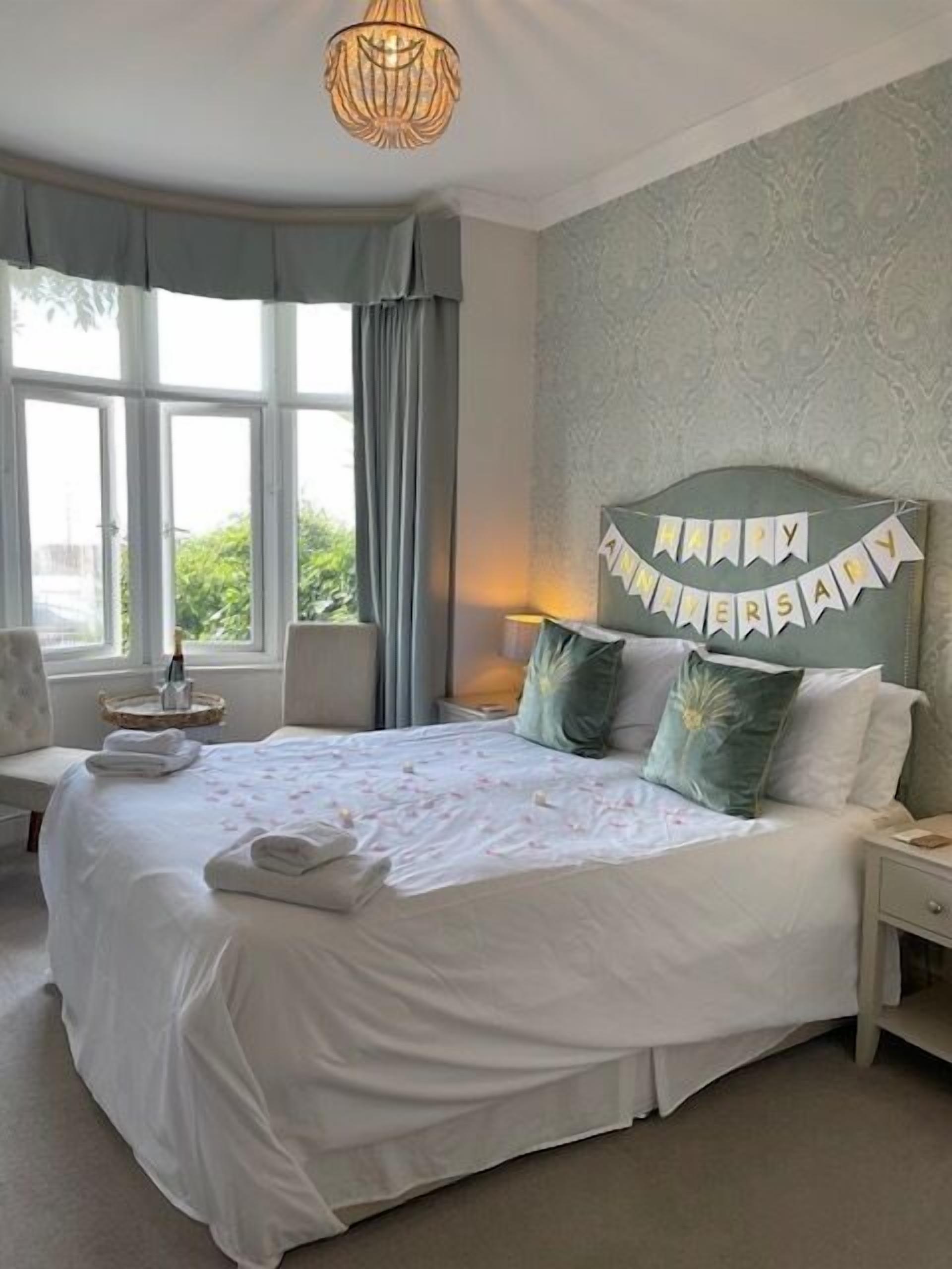A neatly made bed with a 'Happy Anniversary' banner, a bedside lamp, and a view of greenery through the window.