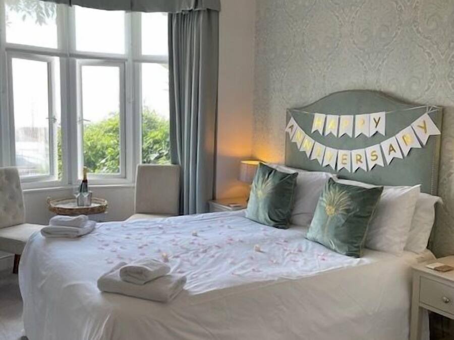 A neatly made bed with a 'Happy Anniversary' banner, a bedside lamp, and a view of greenery through the window.