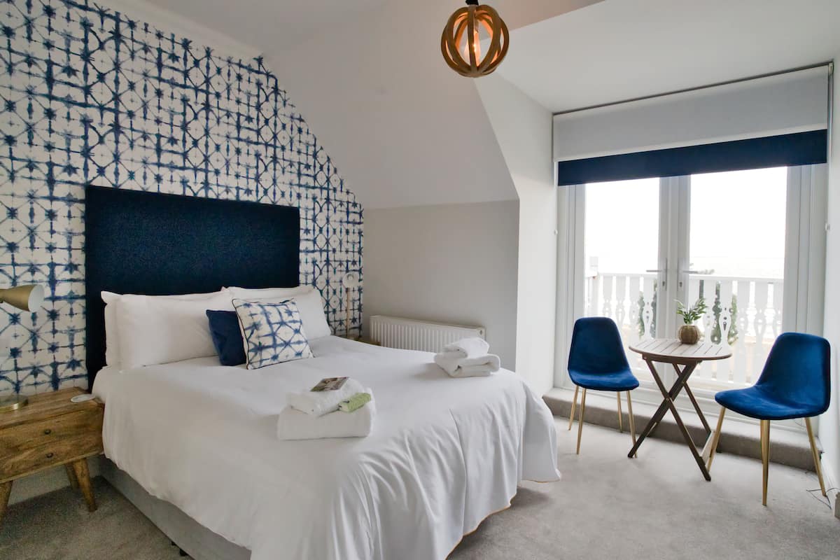 A hotel room with a large bed, a nightstand, a small table with chairs, and a view of the outside through large windows.