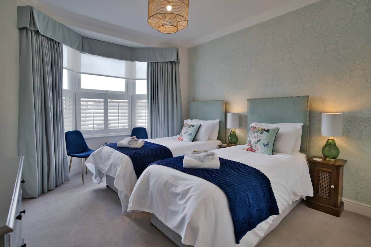 A hotel room with two beds, a blue chair, a small table with a lamp, and a window with curtains.