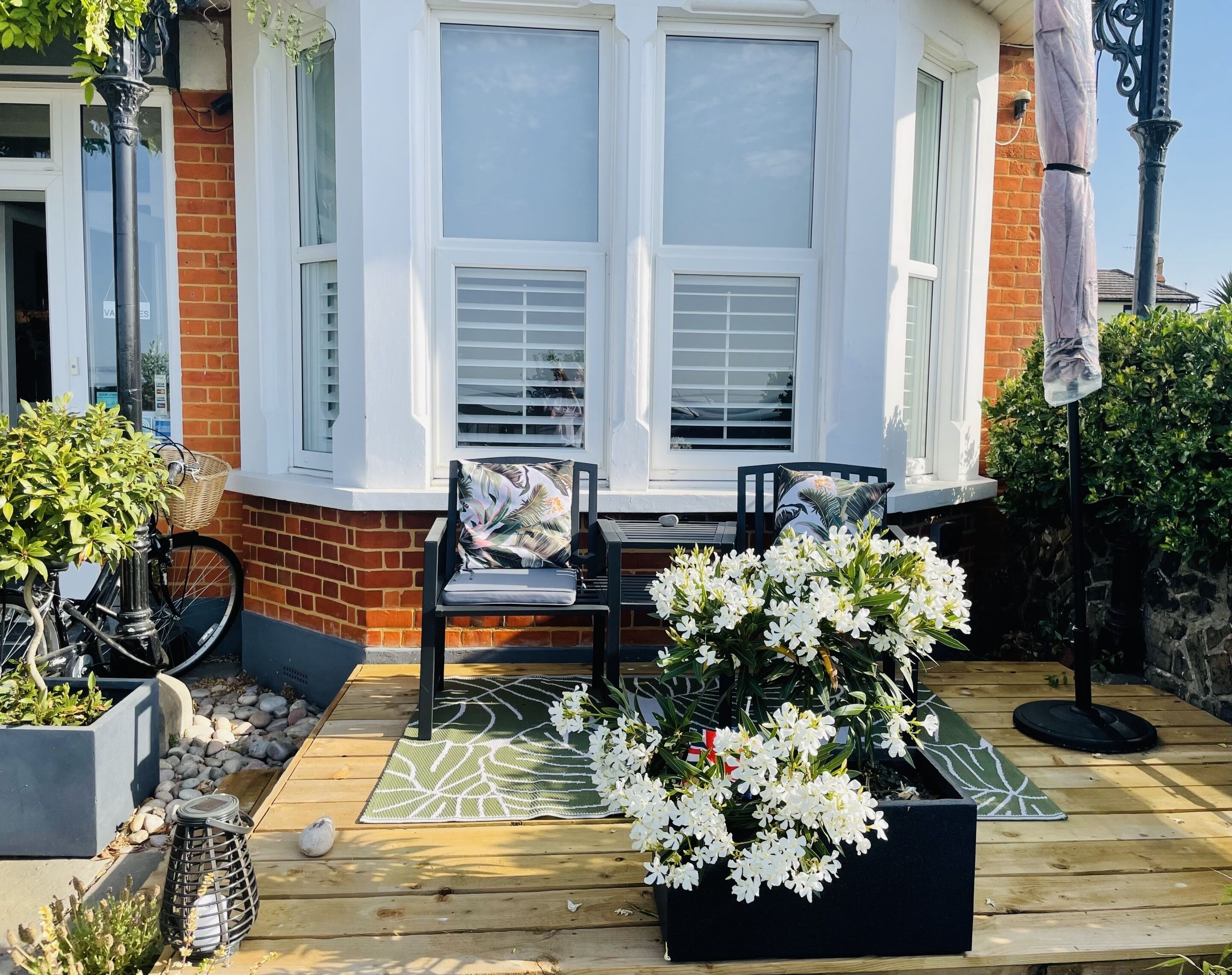 Een houten terras met een erker, twee stoelen, een tafel en een potplant met witte bloemen.