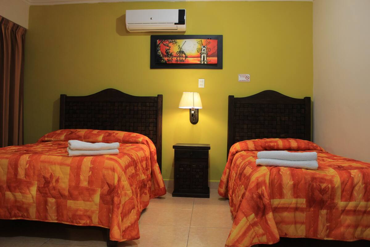A room with two beds, each with a patterned bedspread and a folded towel. There is a wall-mounted air conditioner and a framed picture above the beds.