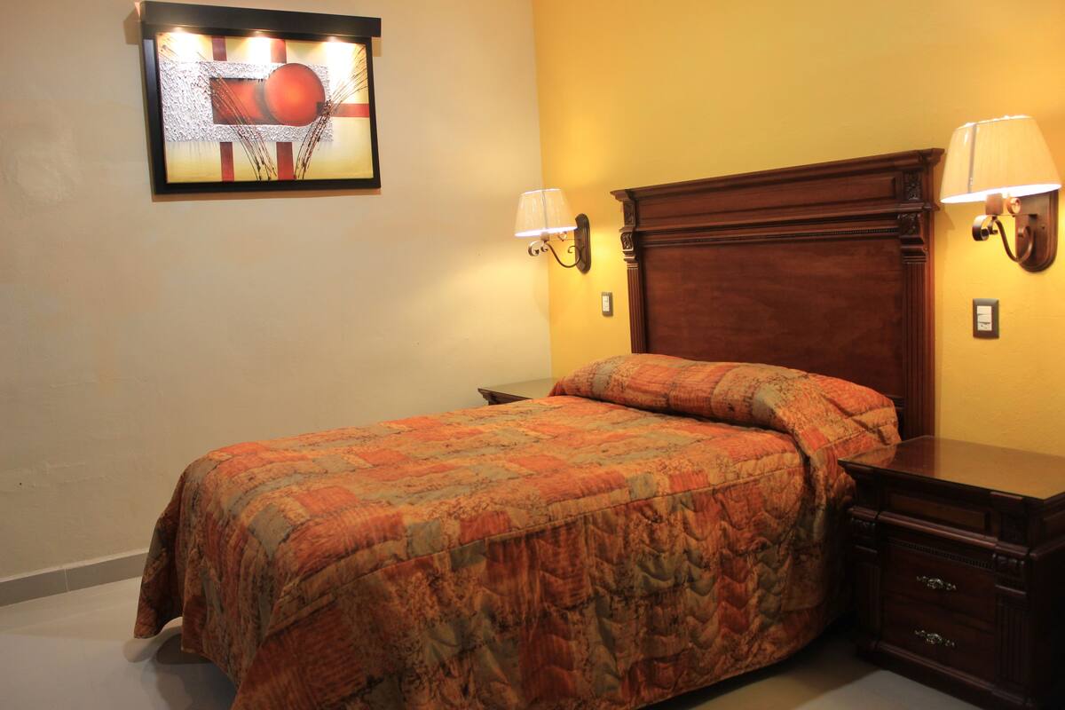 A bedroom with a wooden headboard, two bedside lamps, a bed with a patterned bedspread, a nightstand, and a framed picture on the wall.