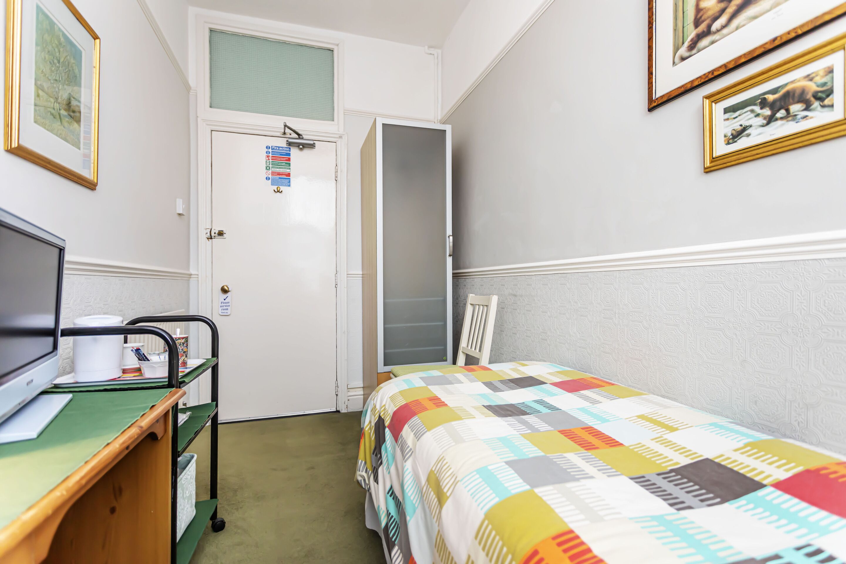 A bedroom with a bed, a desk with a computer, a chair, and a door with a window.
