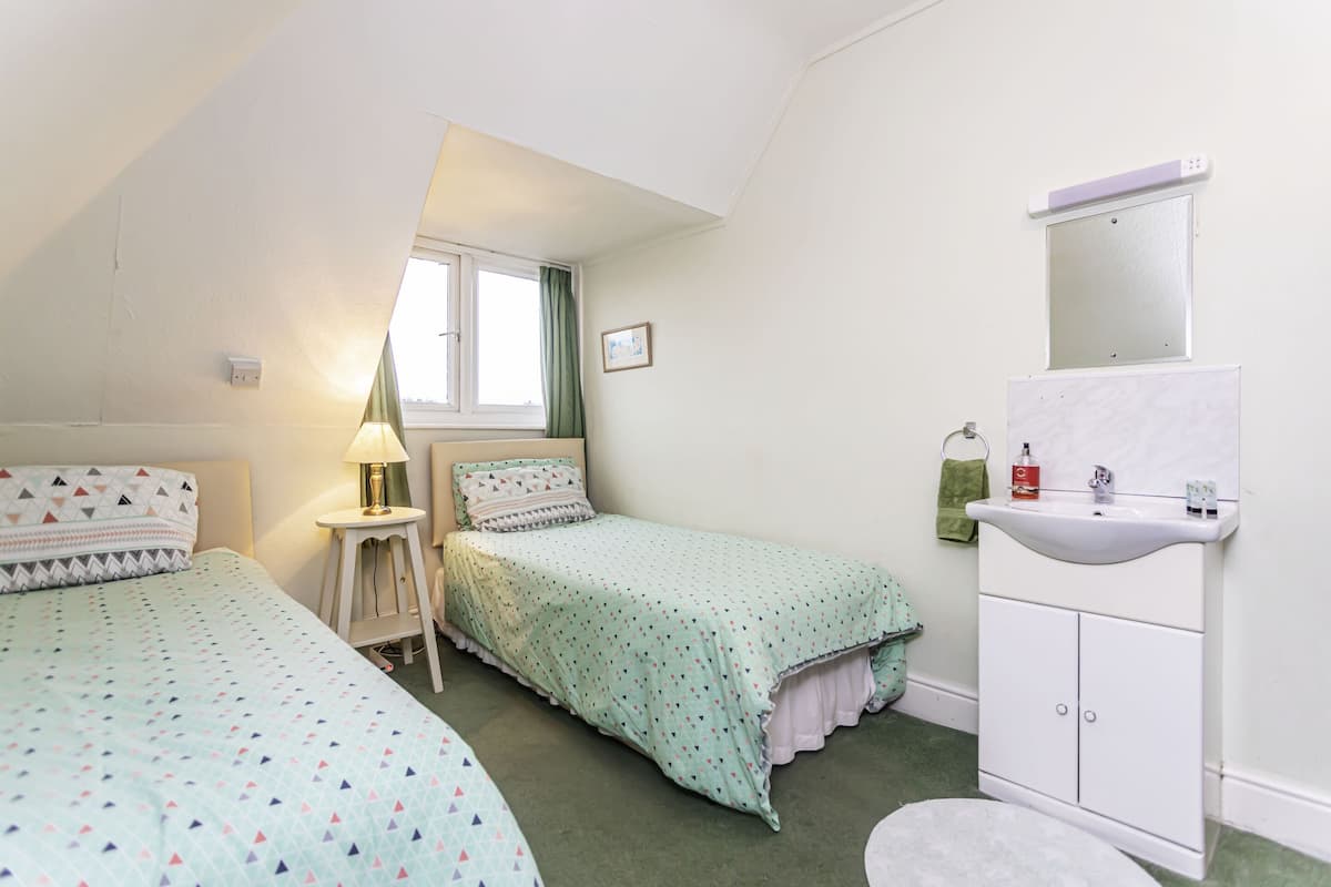 A bedroom with two beds, a window, a bedside table with a lamp, a small cabinet, and a sink.