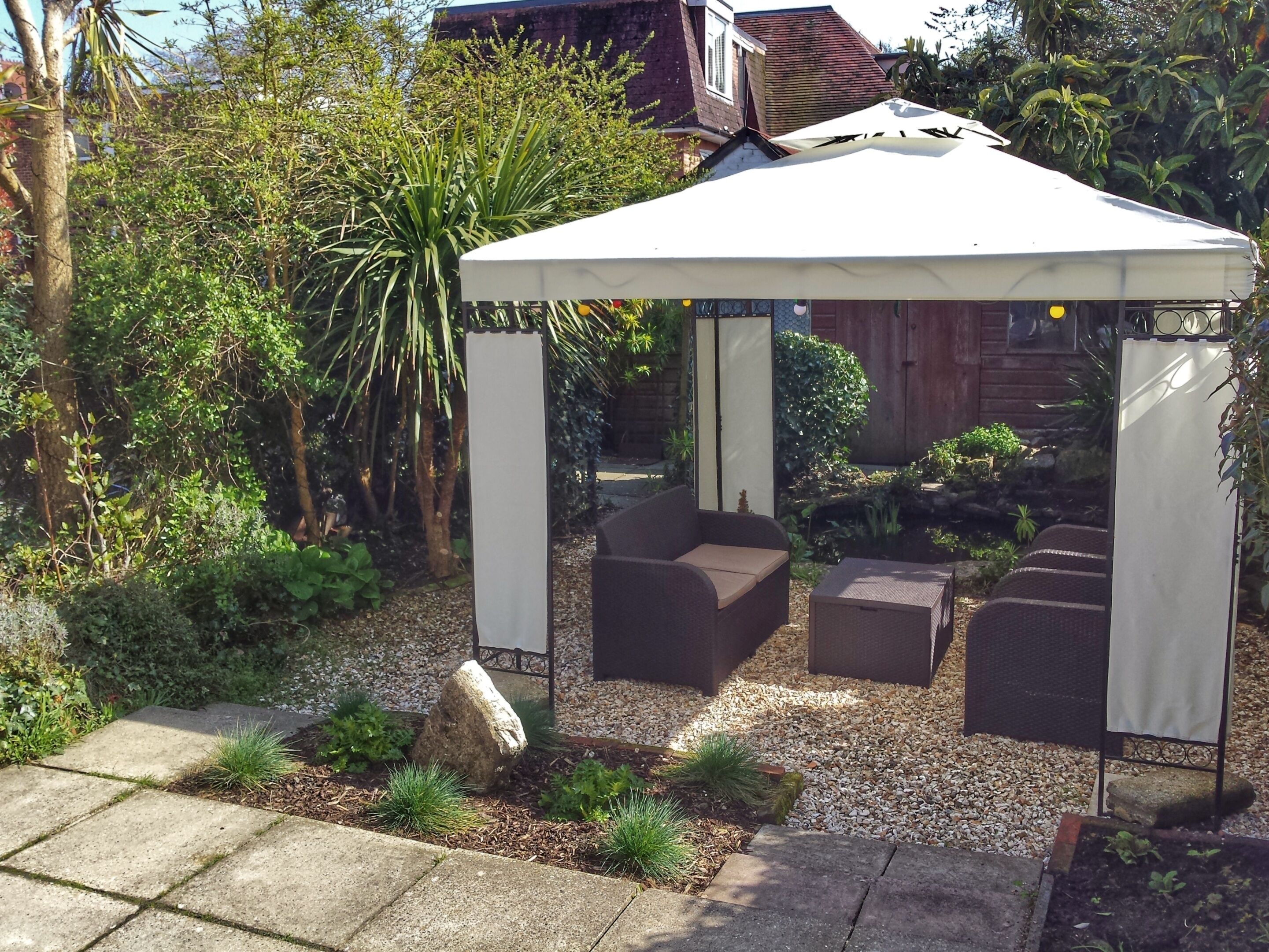 A covered outdoor seating area with a white canopy, surrounded by a garden with various plants and a stone pathway.