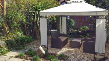 A covered outdoor seating area with a white canopy, surrounded by a garden with various plants and a stone pathway.