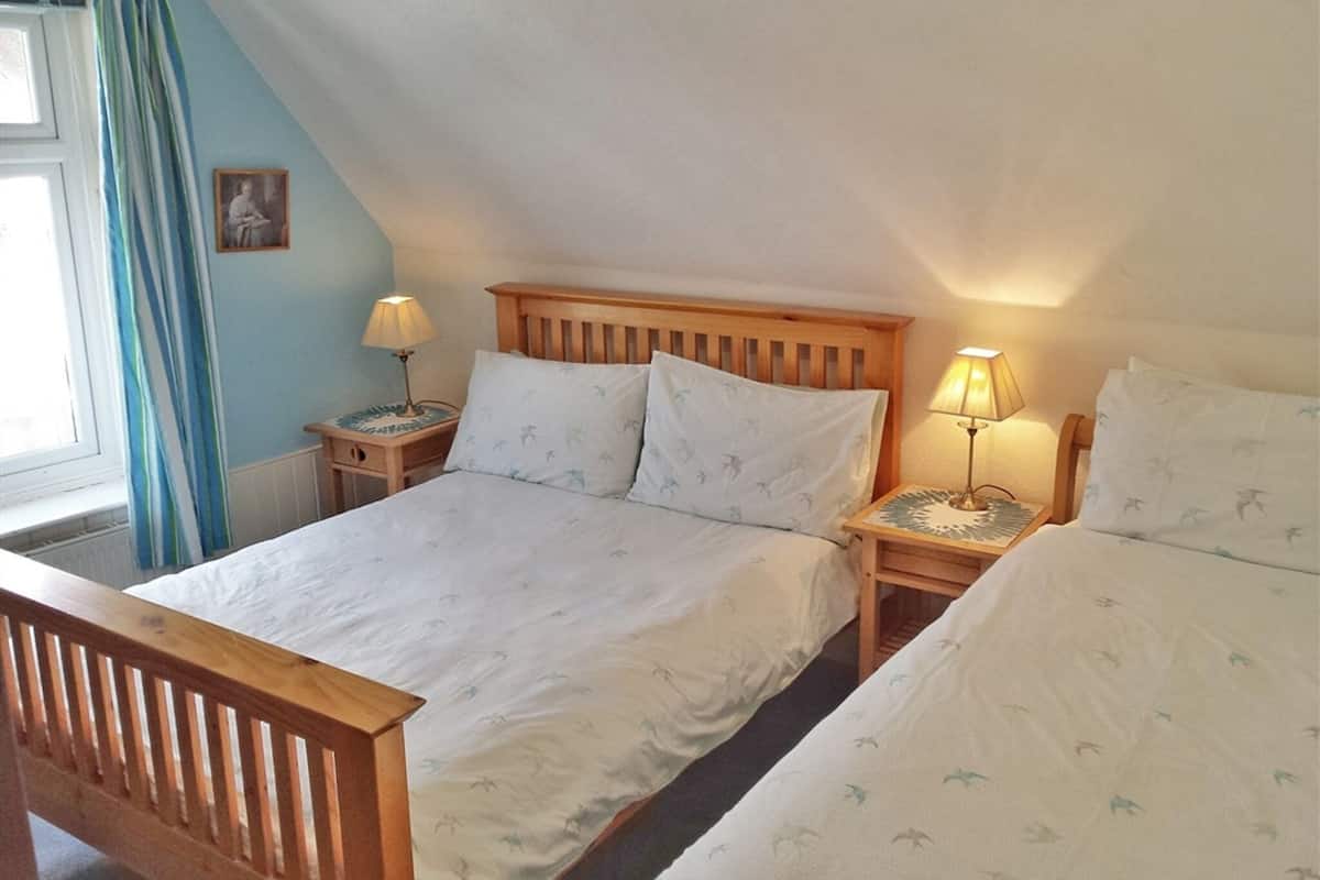 A bedroom with two single beds, wooden headboards, bedside tables with lamps, a window with curtains, and a small framed picture on the wall.
