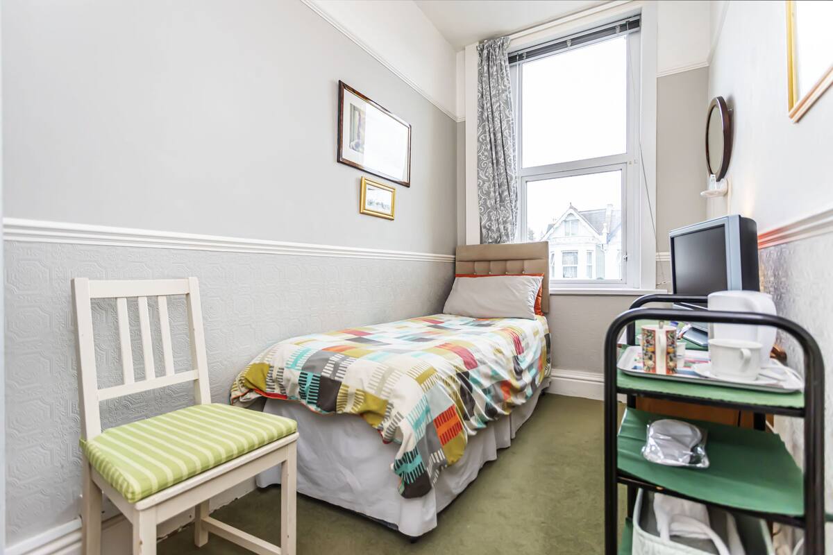 A small, neatly arranged bedroom with a single bed, a chair, a desk with a computer, and a window with curtains.