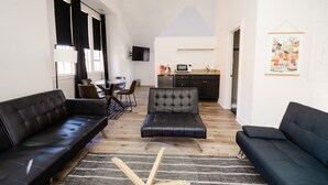 Un salon moderne avec un canapé en cuir noir, un coin repas avec une table et des chaises, une cuisine équipée d’un four à micro-ondes et un téléviseur fixé au mur.