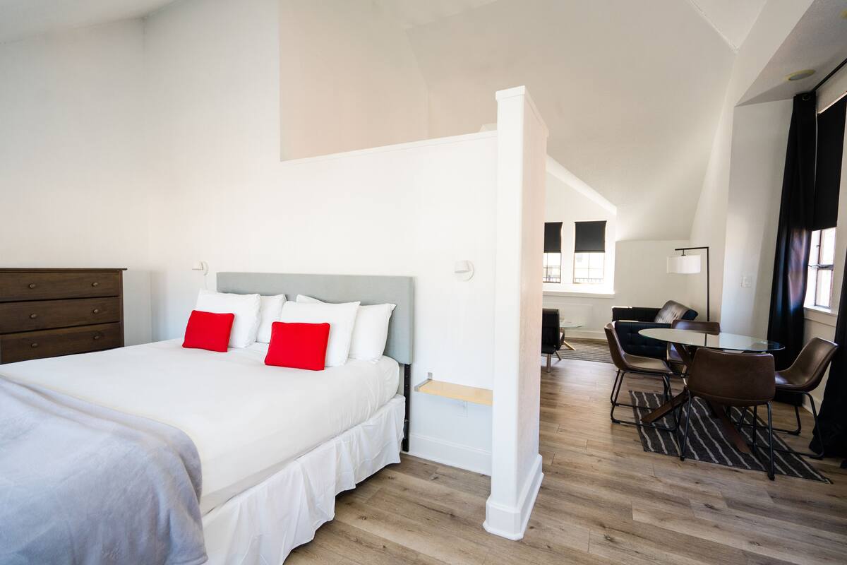 A modern hotel room with a large bed, a desk, and a dining area.